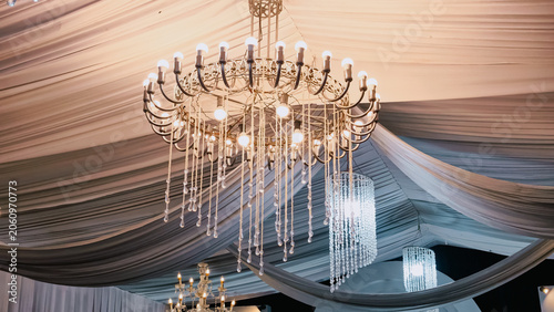 A Magnificent Crystal Chandelier Casts a Luxurious Glow Above the Elegantly Draped White Fabric Ceiling, Creating a Luxurious and Festive Atmosphere for Weddings and Celebrations.