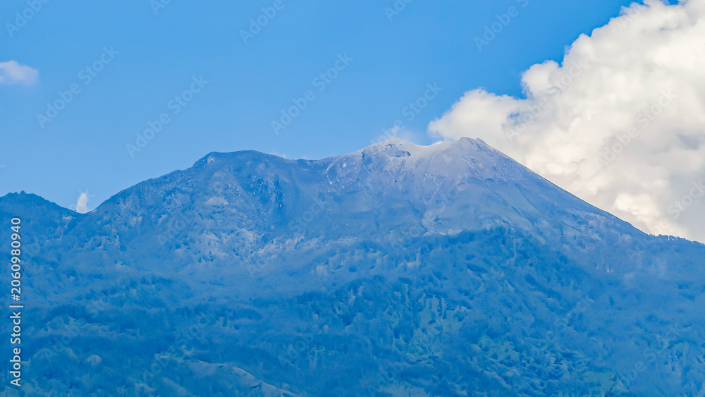 Obraz premium Majestic volcanic mountain peak rising under a clear blue sky with soft white clouds, showcasing the breathtaking grandeur and untouched beauty of nature's powerful landscapes