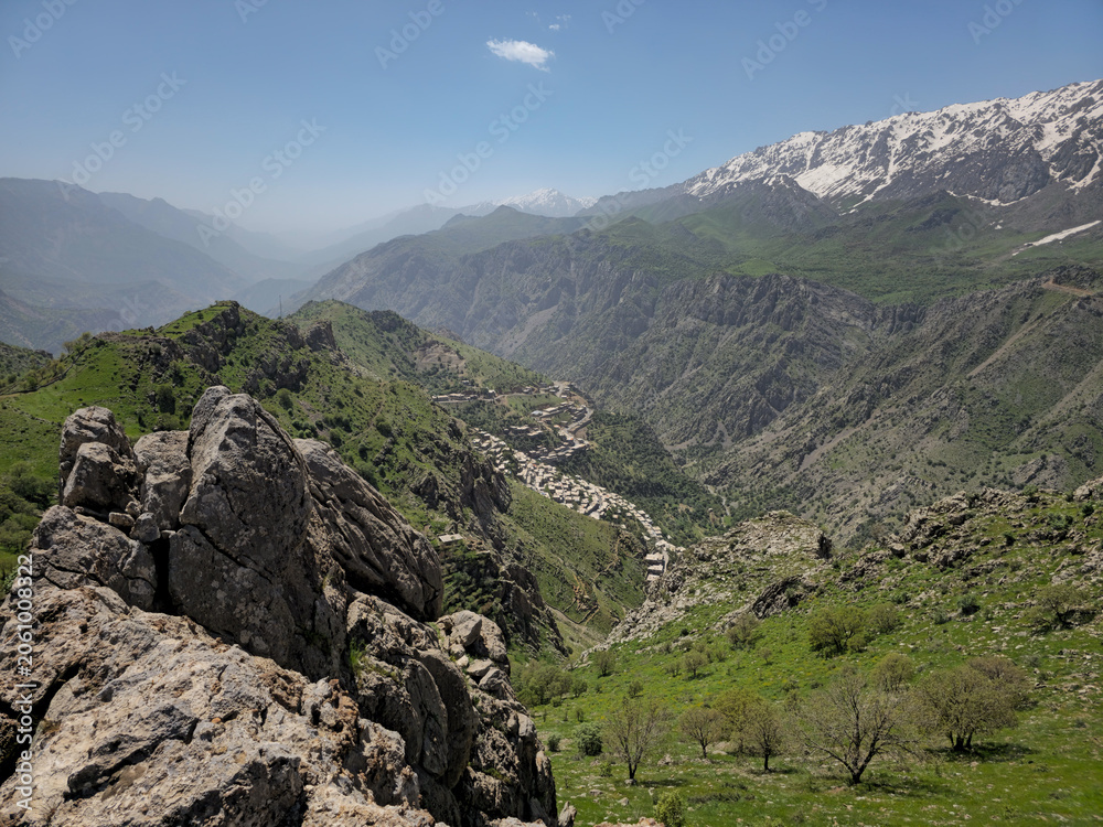 Obraz premium Jagged rocks frame a sweeping spring valley with a nestled mountain village, green slopes, and snow-capped peaks beyond. 📍 Hawraman Takht, Kurdistan, Iran 