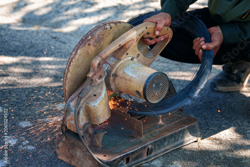 Worker hands cutting roll of steel wire with an electric chop saw machine, bright sparks flying. Industrial work concept.