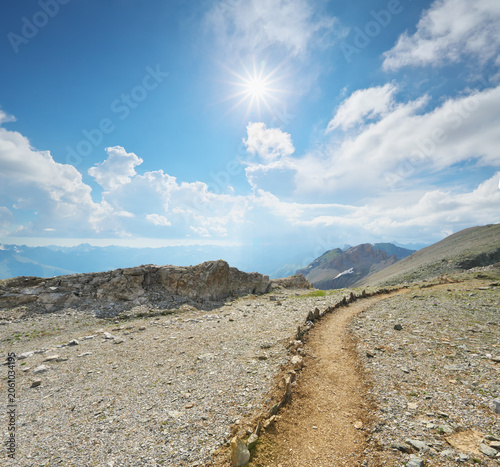 Scenic hiking trail winding through a rocky mountain ridge under a bright sun burst sky. Beautiful alpine landscape.