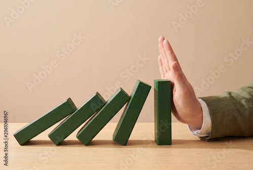 Conceptual hand stopping falling green wooden blocks, interrupting a chain reaction. Minimal clean background for business risk management, problem-solving, and crisis intervention themes.