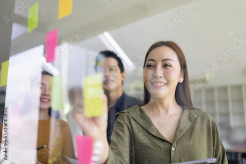 A group of young Asian business colleagues brainstorming and sharing ideas during a meeting, pointing at colorful sticky notes on a glass window in a modern office.