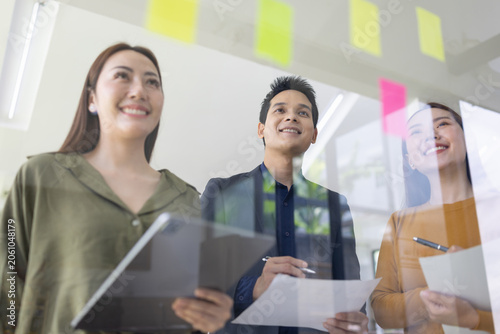 A group of young Asian business colleagues brainstorming and sharing ideas during a meeting, pointing at colorful sticky notes on a glass window in a modern office.