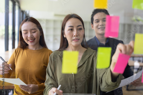 A group of young Asian business colleagues brainstorming and sharing ideas during a meeting, pointing at colorful sticky notes on a glass window in a modern office.