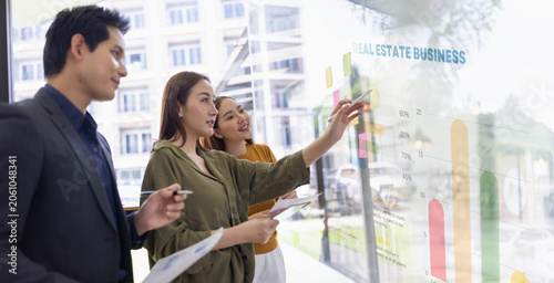 A group of young Asian business colleagues brainstorming and sharing ideas during a meeting, pointing at colorful sticky notes on a glass window in a modern office.