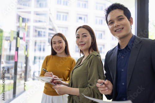 A group of young Asian business colleagues brainstorming and sharing ideas during a meeting, pointing at colorful sticky notes on a glass window in a modern office.