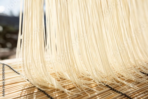Thin white noodles drying on a bamboo blind in soft sunlight.