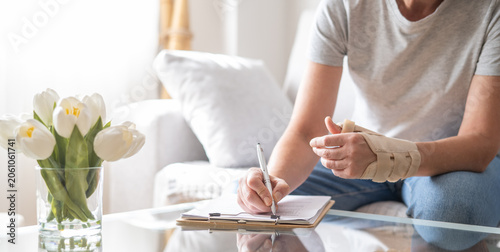 Woman with a medical orthotic wrist splint on her hand, suffering from joint disease, pain, or injury, completing a report for sick leave or treatment while sitting on a couch at home
