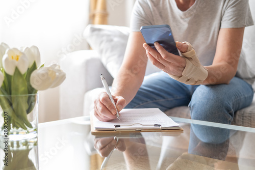 Person wearing an orthotic splint on hand and wrist, holding a smartphone while writing documents and medical reports for sick leave or insurance claims