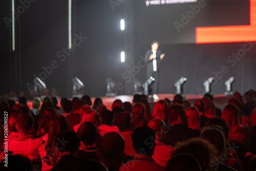 Crowd watching blurred speaker on stage during live conference event, dramatic red lighting creates intense, focused atmosphere for business presentation and public speaking.