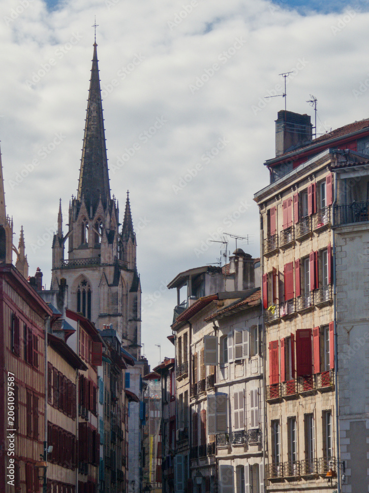 Obraz premium A narrow traditional street in the historic center of Bayonne, France, featuring colorful red shutters and timber-framed houses. The gothic spire of the Bayonne Cathedral rises majestically.