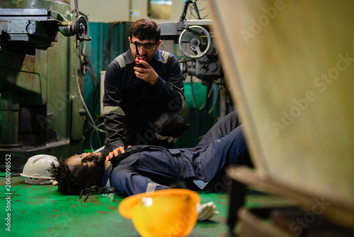 Industrial employee contacting emergency support while monitoring unconscious coworker after workplace incident. Factory safety procedures, emergency response planning, occupational health awareness.