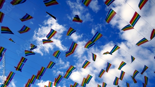 Many colorful rainbow pride flags are flying against a vibrant blue sky with soft white clouds, symbolizing support, diversity, equality, and celebration for the LGBTQ+ community
