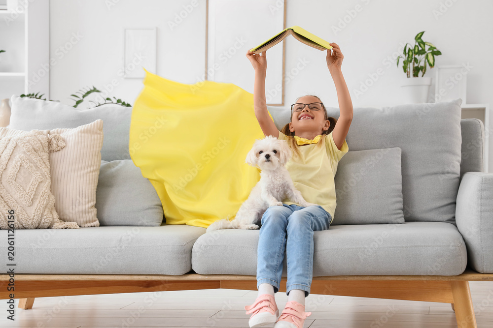 Naklejka premium Cute little happy girl in superhero costume with Maltese dog reading book on sofa at home