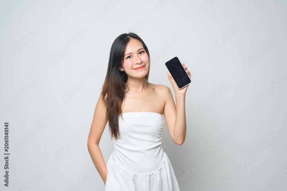 Naklejka premium A young woman in a white dress smiles while holding up a smartphone with a black screen against a plain white background.