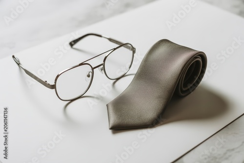 Stylish silver frame eyeglasses resting beside a neatly rolled sophisticated grey silk tie on a professional white surface