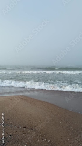 Waves crash gently on sandy shore with misty atmosphere, serene coastal scene in slow-motion