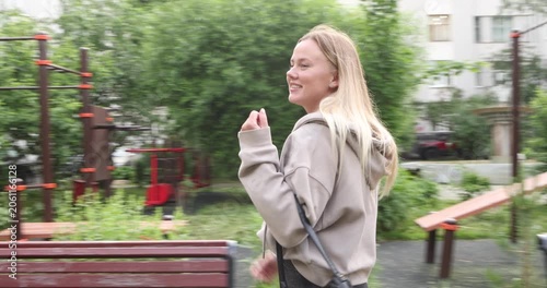 Cheerful young blonde woman wearing a hoodie and wide-leg jeans happily spinning and dancing with a smile while walking along a path in a beautiful, green city park during the daytime