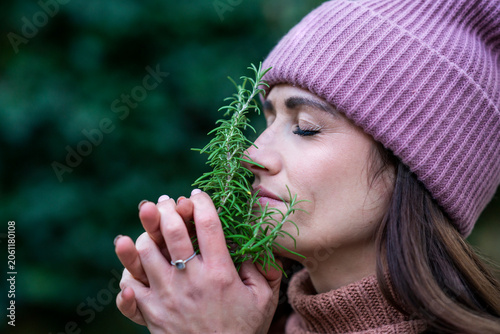 Portrait of beautiful woman wearing pink knit hat smelling fresh rosemary with closed eyes