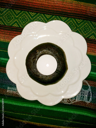 A dark, glossy ring‑shaped sweet sits on a white plate placed over a colourful woven textile with geometric patterns