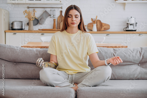 Young woman with a bionic prosthetic arm is sitting on a sofa in a living room, meditating with her eyes closed, fostering wellness and focusing on her mental health