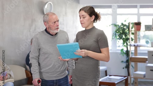 Positive polite young secretary or social worker talking to aged man visitor in reception area in background queue of people 