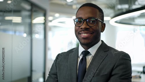 Young african-american businessman in charcoal suit, white shirt, black-rimmed glasses, smiling confidently, arms crossed in modern corporate office with blurred colleagues. concept of professional