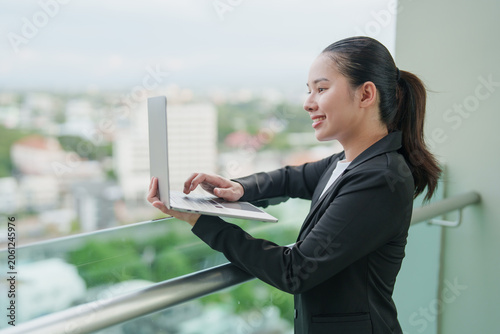 Fotografija Asian businesswoman working remotely on a laptop, standing happily outdoors on a