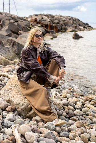 Stylish woman sitting on rocky beach beside calm sea in relaxed autumn fashion lifestyle portrait