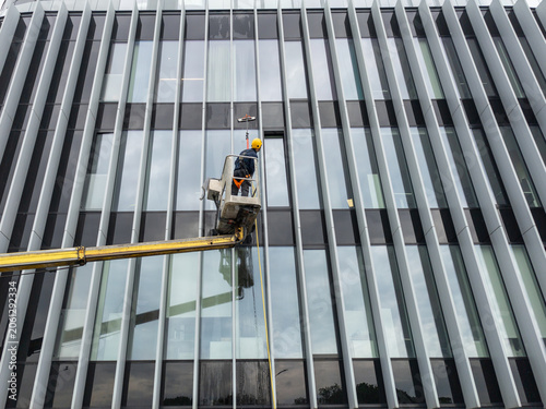 Professional window washer cleaning glass facade of modern office building using cherry picker crane, maintenance worker washing windows on high rise skyscraper