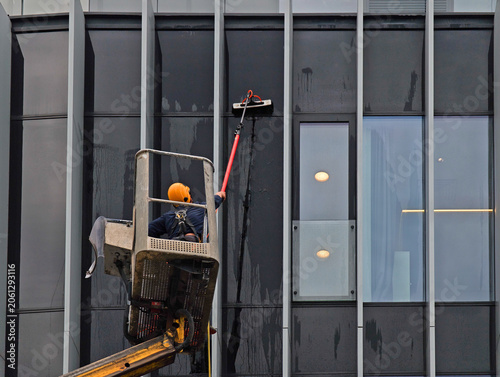 Professional window washer cleaning glass facade of modern office building using telescopic pole from a cherry picker platform, industrial maintenance service