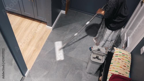 A teenager cleans gray tile with a flat mop beside a gray bucket during an indoor home cleaning routine.