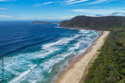 Aerial scene capturing Seven Mile Beach near Forster, NSW, Australia, stretching towards Booti Hill beneath a cloudy blue sky. The expansive sandy shoreline, rolling ocean waves and surrounds.