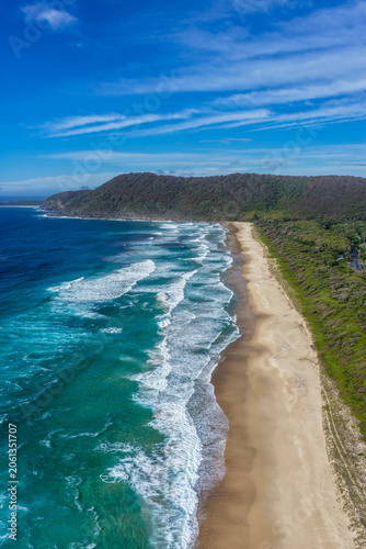 Aerial vertical scene capturing Seven Mile Beach near Forster, NSW, Australia, stretching towards Booti Hill beneath a cloudy blue sky. The expansive sandy shoreline, rolling ocean waves.