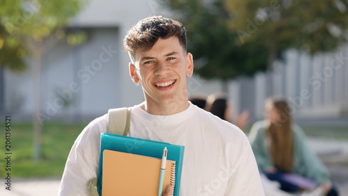 Smiling young man student holding notebooks and backpack on university campus, ready for classes. Peers in background