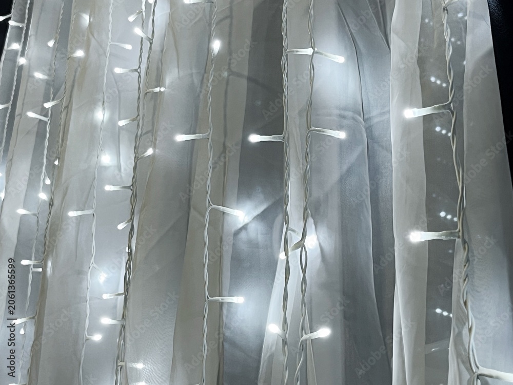 Naklejka premium A string of white LED lights arranged in vertical strands, elegantly draped over translucent white tulle fabric, creating a soft, glowing effect