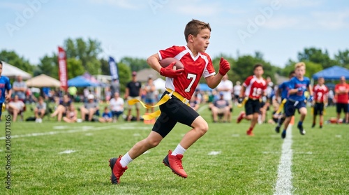 Young white boy running with football during a flag football game on green field. Youth sports competition and outdoor team play at a local athletic field.