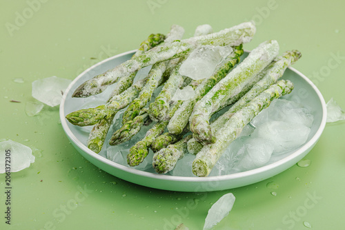 Frozen green asparagus spears covered in hoarfrost with ice cubes on a green background