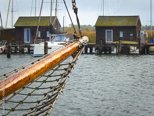 Foto In the harbor of Gager on the island of Rügen, the wooden bowsprit of a sailing ship juts out towards the characteristic boathouses, which stand on wooden stilts in the water