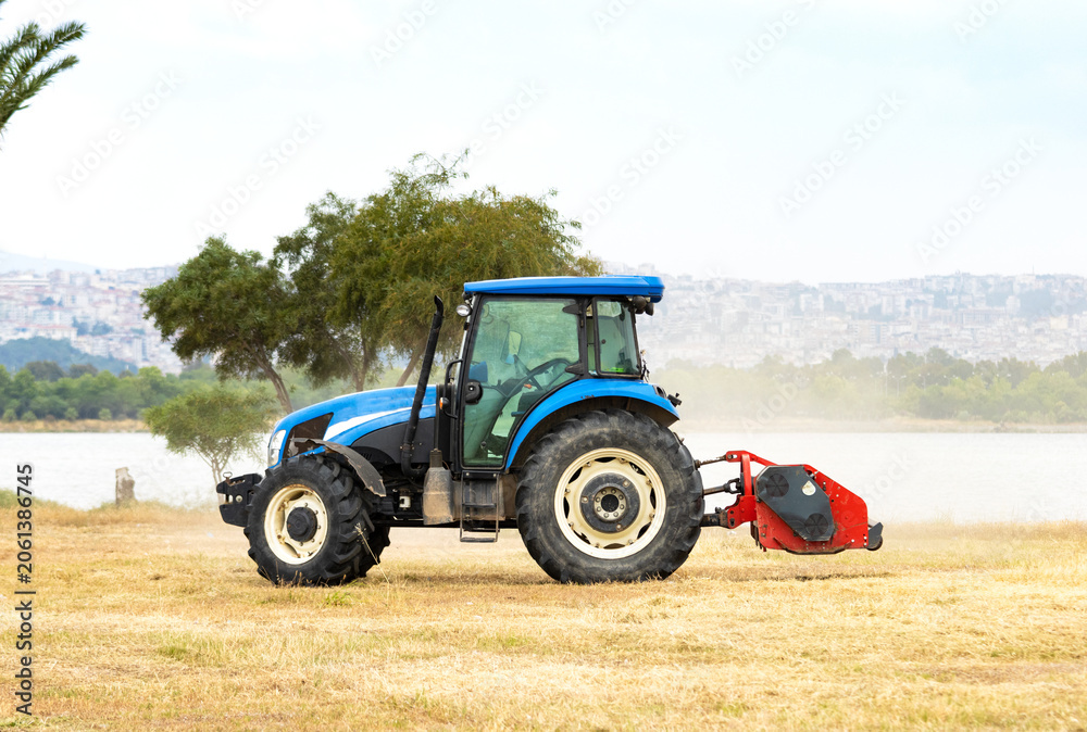 Obraz premium Modern blue agricultural tractor with mechanical equipment working on a dry grass field near a lake side with a distant city skyline under cloudy sky.