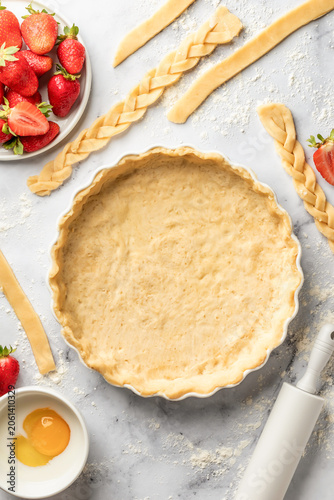 Process of making strawberry shortbread pie. Raw rolled dough in ceramic baking form on marble background with strips of dough around. Home baking culinary concept, recipe. Flat lay