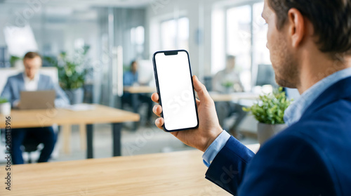 Hand holding mobile phone with blank white screen mockup, professional man in modern office, using smartphone for business tasks, mobile app collaboration or corporate workflow.
