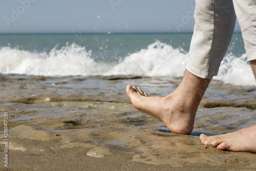 Bare feet walking on wet sandy beach with ocean waves at shoreline, person wearing rolled-up pants.