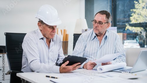 Two senior men review building plans in an office, using a laptop and documents. Safety helmets and blueprints suggest engineering, construction planning, teamwork and project discussion.