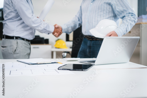 Фотография Two men shake hands in a bright office, one wearing a hard hat, suggesting a successful construction meeting, contract agreement, or project partnership
