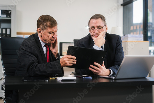 Two business men in suits sit at an office desk looking stressed and frustrated during a difficult meeting, with a laptop and paperwork in front of them.