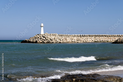 Картината върху платно White lighthouse on rocky breakwater with sea waves crashing in foreground, turquoise water and blue sky, peaceful coastal seascape on sunny day