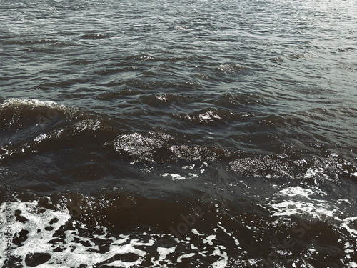 sea waves dark brackish waves ripple across a coastal bay, with foamy water, sunlight reflections, and textured surface patterns creating a natural abstract seascape ocean background