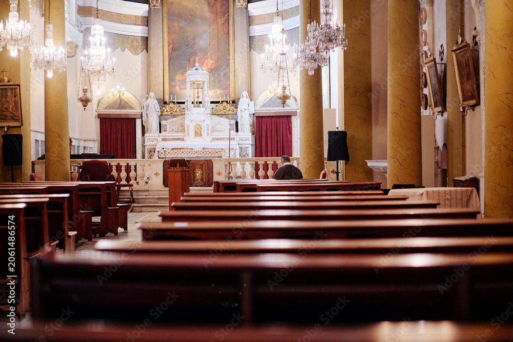 Naklejka premium Quiet church interior with wooden pews, chandeliers, altar, religious statues and a seated worshipper in warm light.